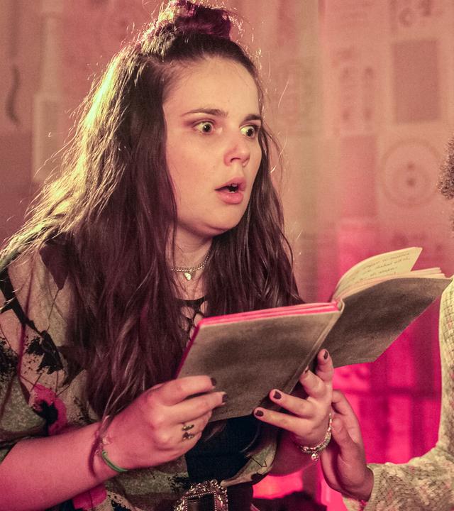 Amy schaut erschrocken und hält ein Buch aufgeschlagen in beiden Händen. Neben ihr steht Cece und schaut total ängstlich. Beide sind in einem fremden Raum mit gelblichem, unheimlichem Licht.