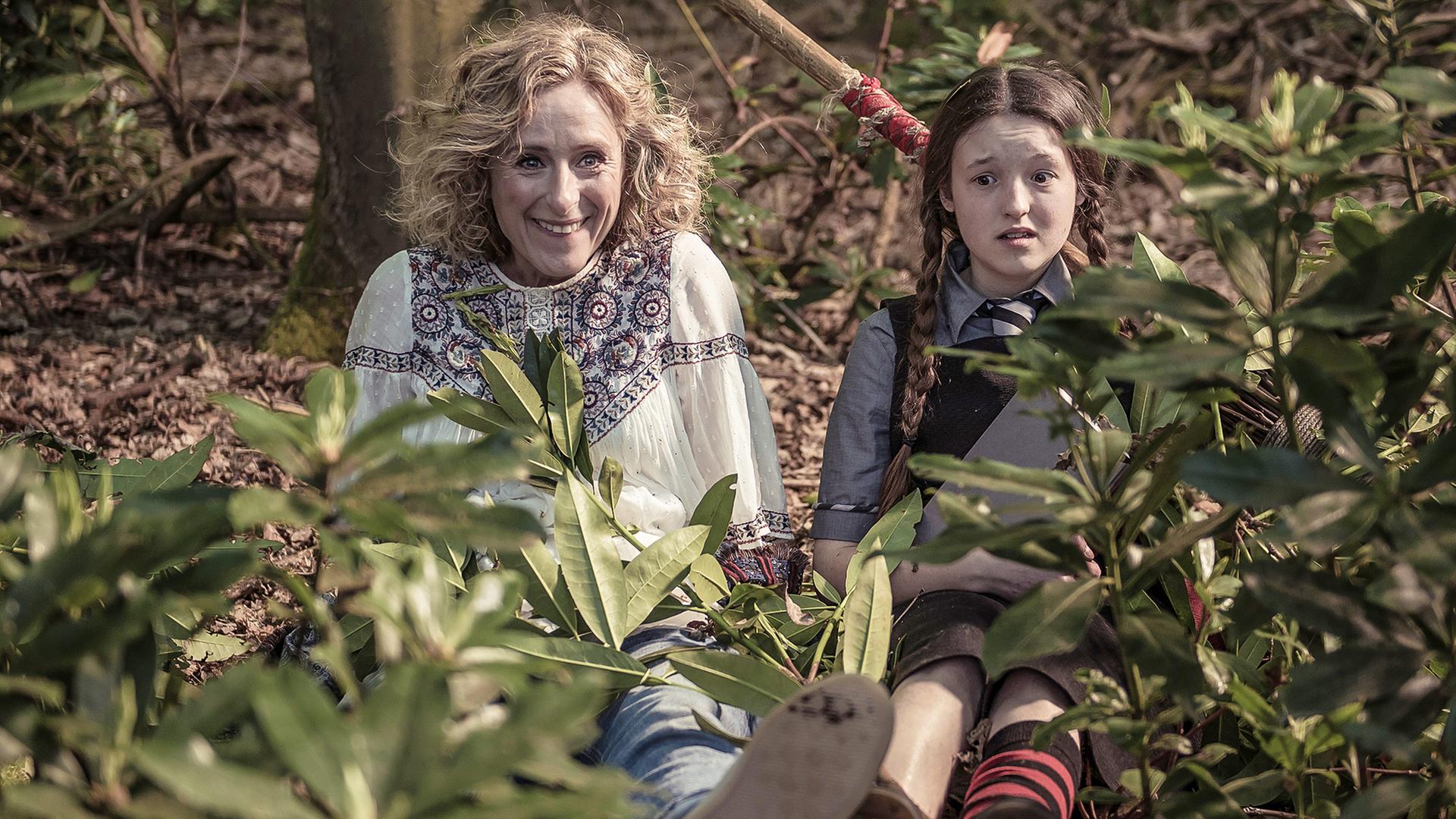 Mildred und ihre Mutter sitzen auf dem Waldboden. Julie lächelt und Mildred schaut verwirrt.