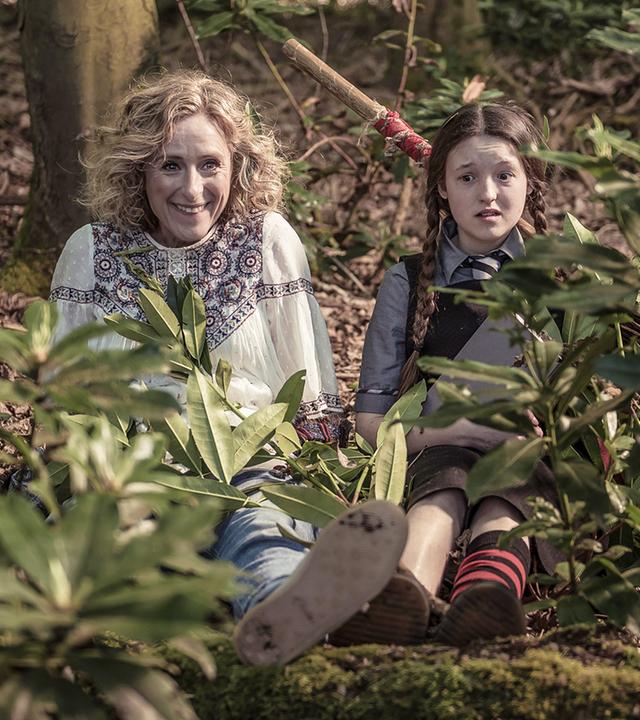 Mildred und ihre Mutter sitzen auf dem Waldboden. Julie lächelt und Mildred schaut verwirrt.