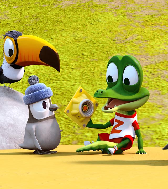 Krokodil Zacki hält freudestrahlend ein Ersatzteil für die Bahn in seiner Hand. Die Pinguine Kurti und Jonathan und der Tukan Toni schauen Zacki dabei interessiert zu.
