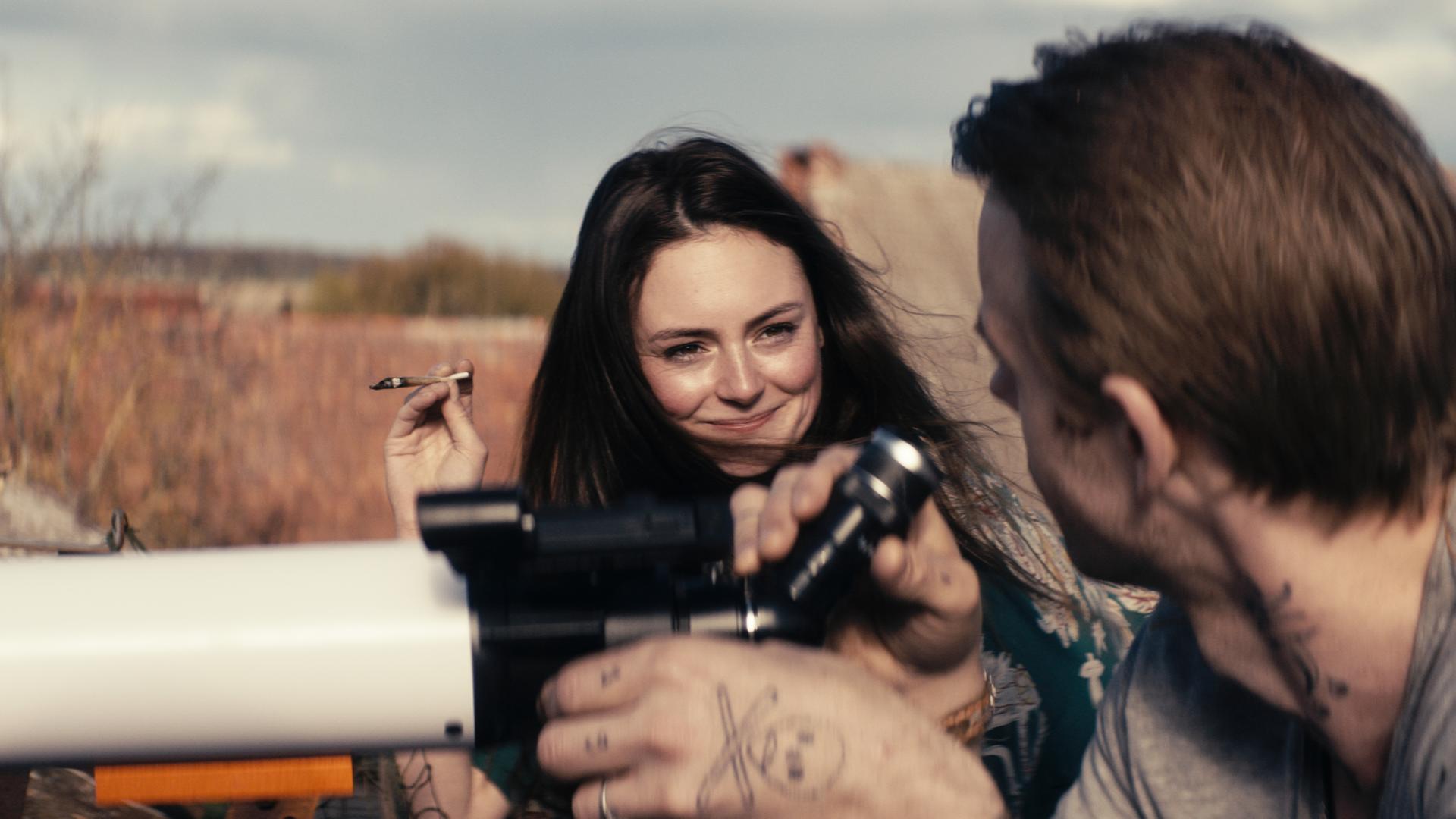 Ella (Lea van Acken) hält eine Zigarette in der Hand. Sie sitzt neben Jan (Mads Hjulmand), der etwas durch ein Fernrohr beobachtet.