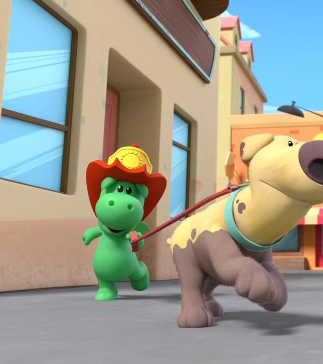 Grisu, der kleine grüne Drache mit dem roten Feuerwehrhelm, hat den Hund Dinki an der Leine. Beide rennen sie durch eine Straße von Drachenstadt. Etwas abseits beobachtet sie dabei Stella.