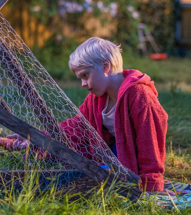 Pelle sitzt draußen im hohen, leicht verwilderten Gras neben einem kleinen Außengehege aus Holz und Drahtgitter. Vor ihm im Gehege sitzt ein dunkles Kaninchen, zu dem er sich aufmerksam und ruhig hinbeugt.