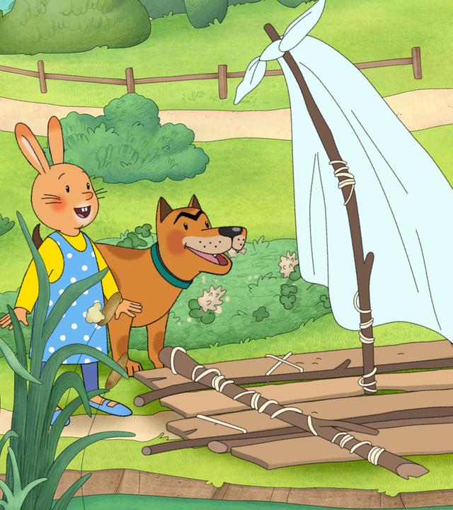 Karlchen, der Kaninchenjunge, und seine Freundin Monika mit ihrem Hund Bello stehen bei einem selbstgebauten Floß mit einem weißen Segel auf einer Wiese. 