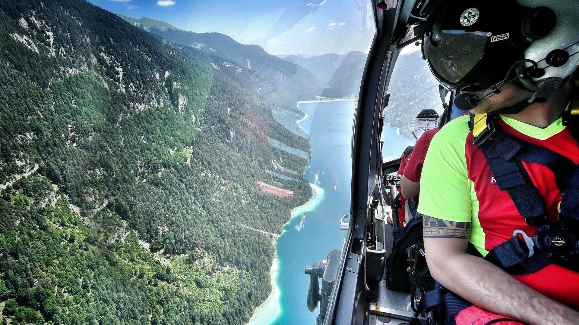 Ein Flugretter schaut aus dem Fenster des Heli. Unterhalb liegt ein malerischer Gebirgssee. 