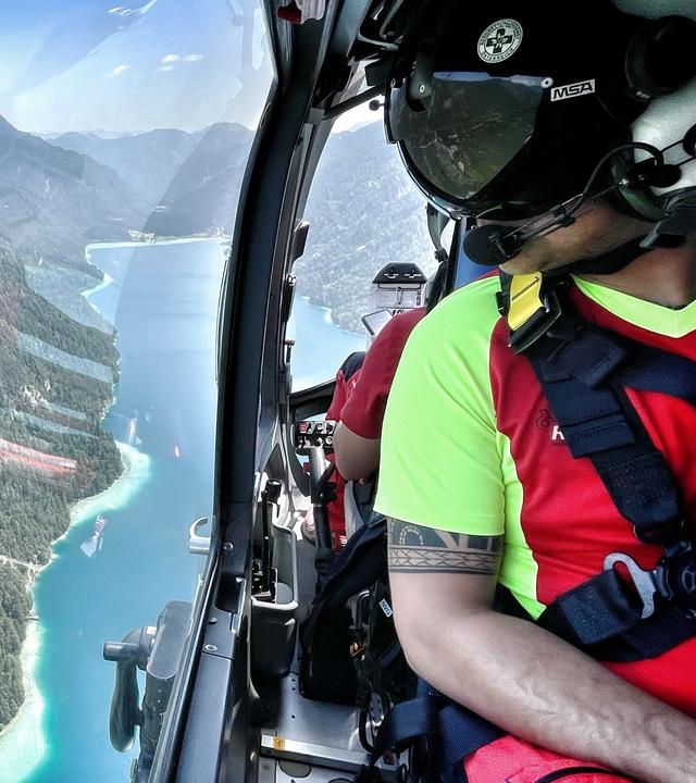 Ein Flugretter schaut aus dem Fenster des Heli. Unterhalb liegt ein malerischer Gebirgssee. 
