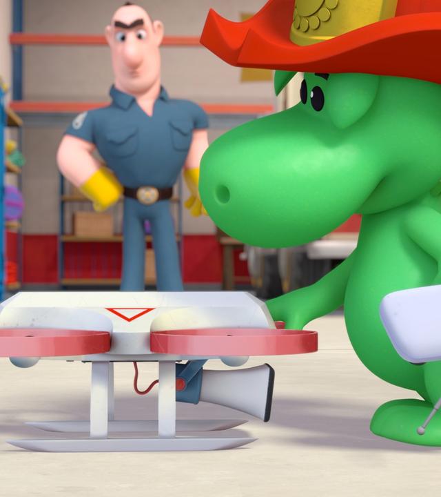 Grisu, der kleine Drache, steht vor der Feuerwache. Er schaut sich eine Drohne ganz genau an. In seiner Hand hält er eine Fernbedienung. Im Hintergrund schauen die Feuerwehrleute Olivia und David dem Drachen zu.