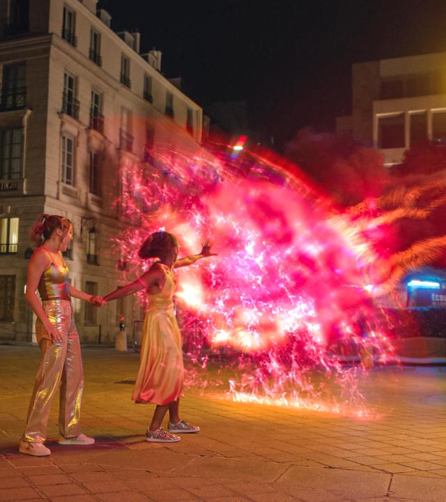 Abends auf einer Straße in Paris: Jade und Cece werden von dunkler Mystic-Magie angegriffen. Cece wehrt diese mit Wizen-Kräften ab. Ein rot glühender Schutzwall baut sich vor ihnen auf. Jade steht hinter Cece und hält deren Hand fest.