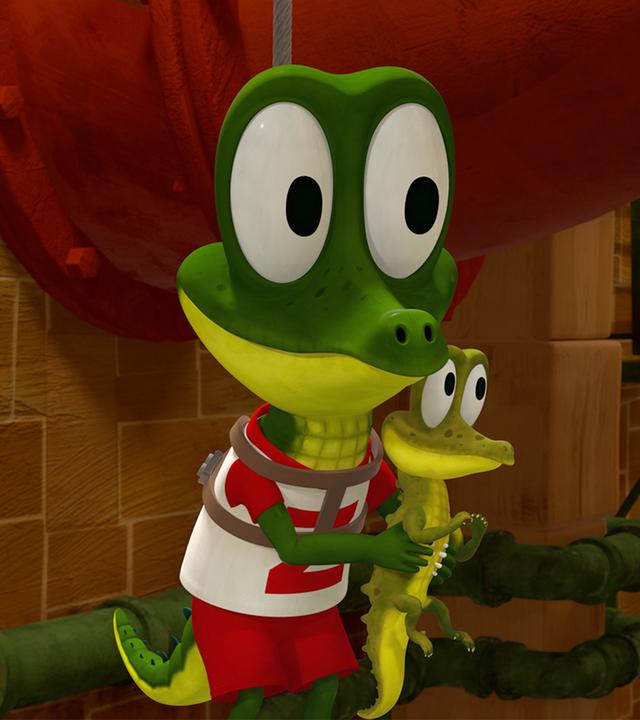 Krokodil Zacki hält ein ganz kleines Krokodil in seinen Händen, welches Giraffe Bruno freudig begutachtet.