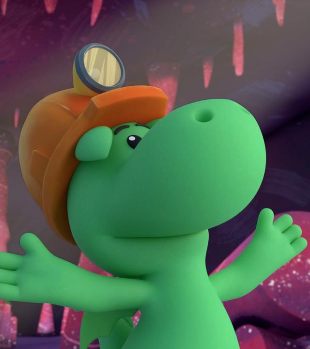 Grisu, der kleine Drache, und seine Freundin Stella stehen in einer Höhle mit rosafarbenen Steinen. Sie tragen Helme mit Stirnlampen. Grisu macht mit den Händen eine ausladenden Geste.