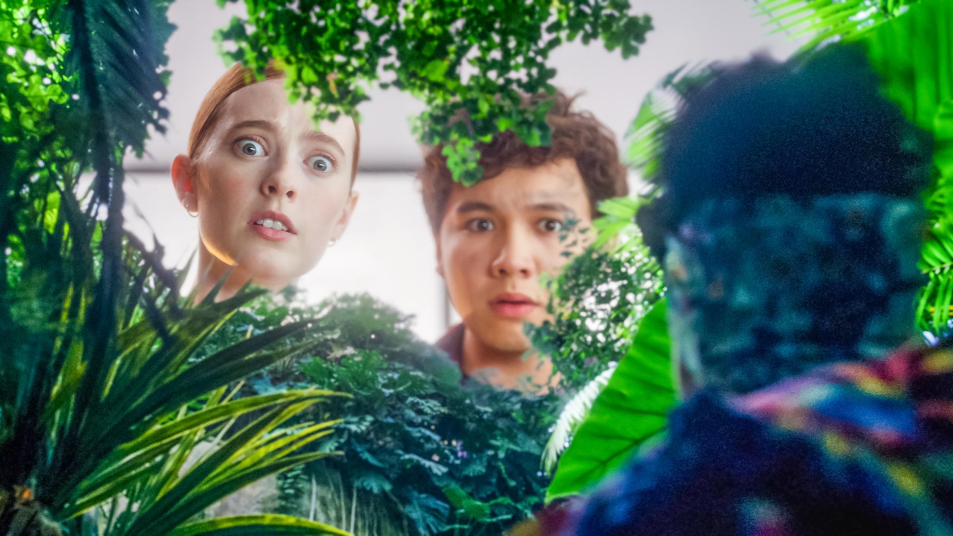 Mia und Bash blicken erschrocken von oben durch ganz viel Grün in ein Gewächshaus. Rechts und nur von hinten in Umrissen zu sehen ist Cece, die ganz klein geschrumpft inmitten des Grüns steht.