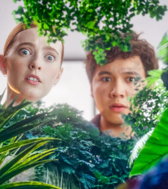 Mia und Bash blicken erschrocken von oben durch ganz viel Grün in ein Gewächshaus. Rechts und nur von hinten in Umrissen zu sehen ist Cece, die ganz klein geschrumpft inmitten des Grüns steht.