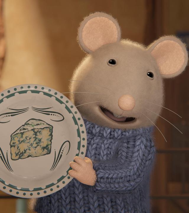 Sam freut sich und hält einen bemalten Teller hoch, auf dem ein Schimmelkäse, ein sogenannter Roquefort, abgebildet ist.