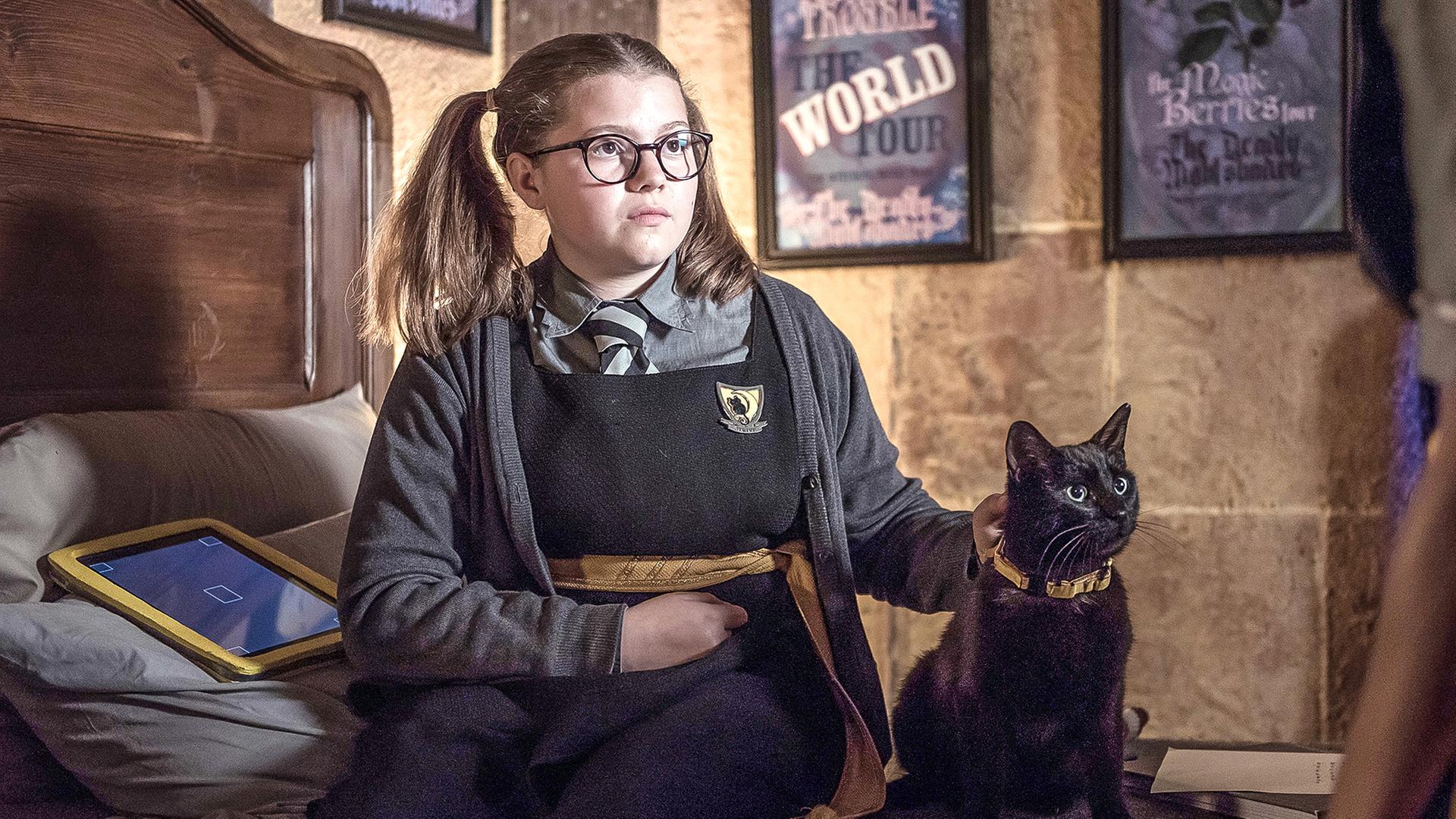 Maude sitzt mit ihrer Katze lächelnd auf ihrem Bett.