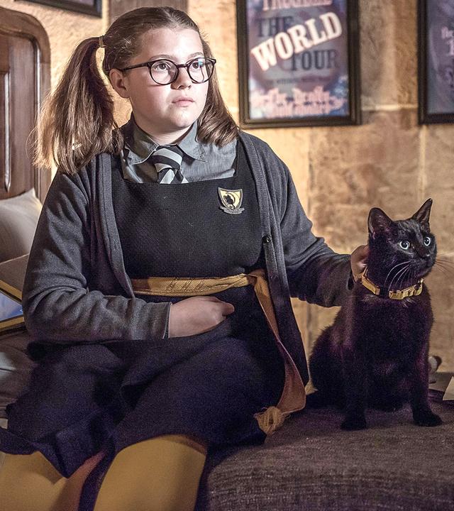 Maude sitzt mit ihrer Katze lächelnd auf ihrem Bett.