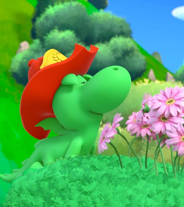 Grisu, der kleine Drache, trägt seinen roten, großen Feuerwehrhelm und steht im Grünen vor rosa Blümchen. Mit seiner Nase riecht er daran.