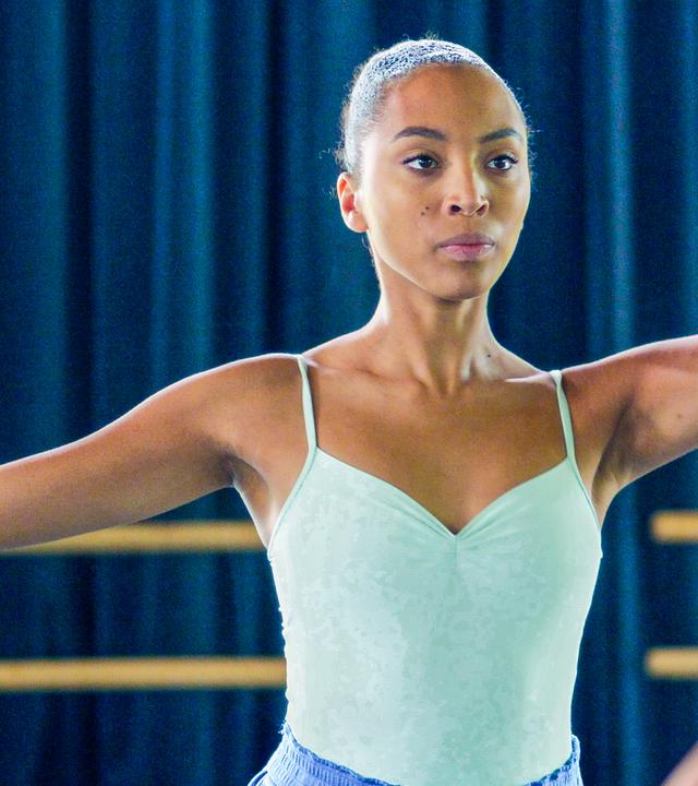 Cece hat ihr Trainingsballett-Outfit an und nimmt eine Tanzpose ein. Ihr linker Arm ist erhoben. Benoit steht ihr gegenüber. Cece schaut angestrengt.