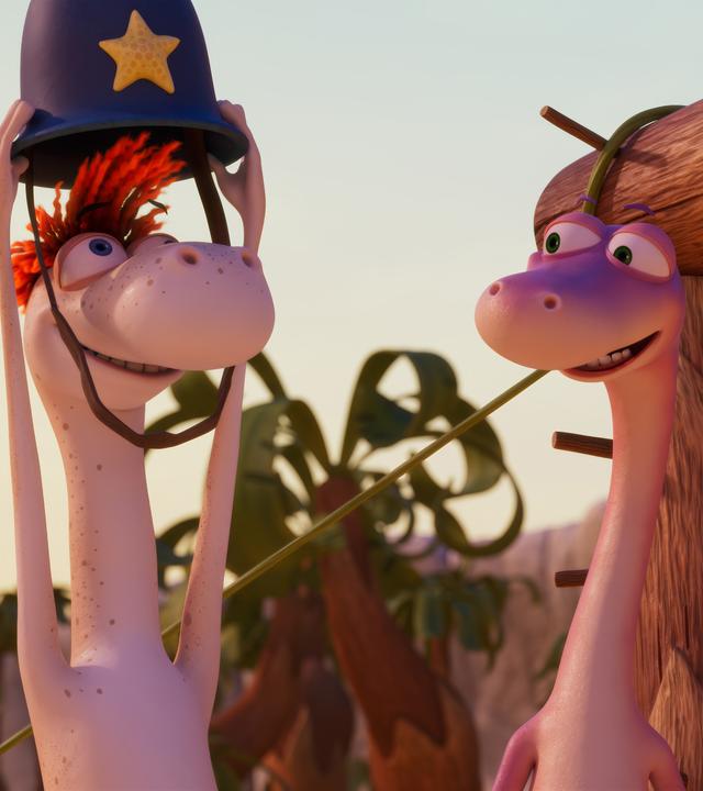 Flint, der Dinosaurier mit den orangefarbenen Wollhaaren, setzt sich gerade den Helm von Wachtmeister Paleowski auf. Er schaut überglücklich. Sein Freund Minus Drei schaut ihm von der Seite aus zu.