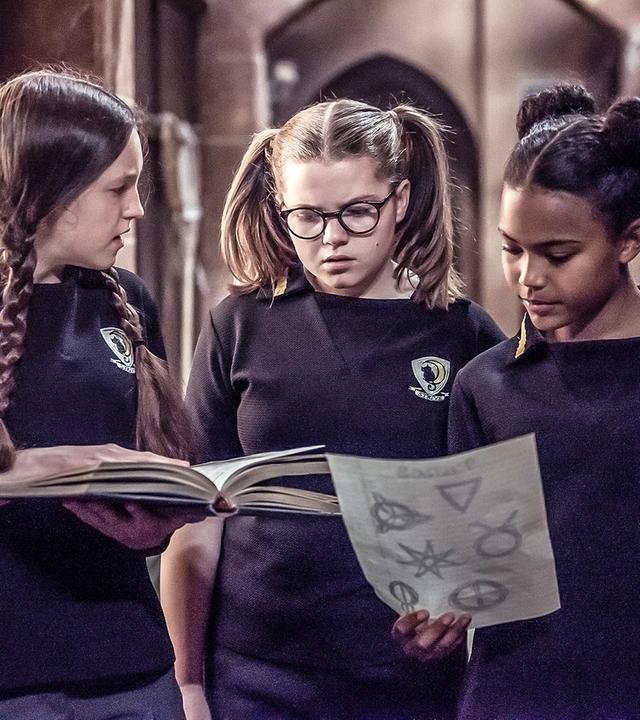 Mildred, Maude und Edith stehen im Schulkorridor und blicken auf ein Blatt mit Zeichnungen und ein Buch.