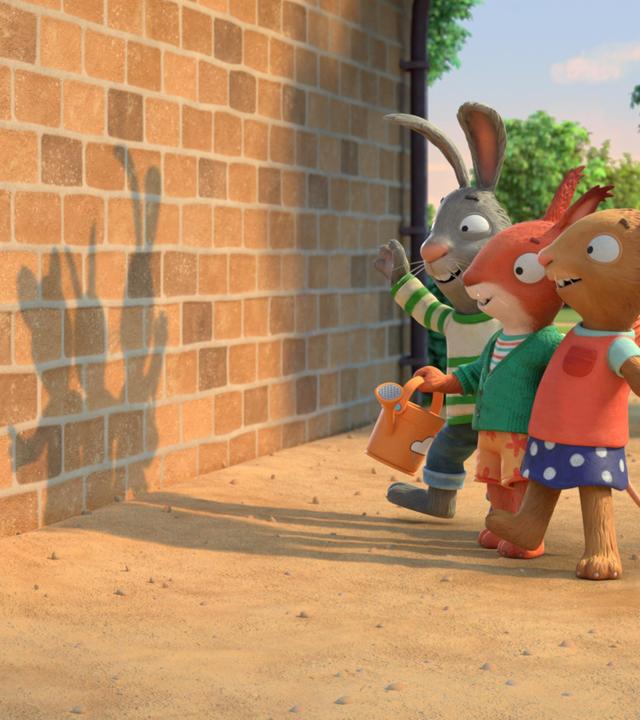 In einer Wohngegend auf einem Weg vor einer Backsteinwand stehen die drei Freunde Pip, das Kaninchen, Posy, die Maus und Jamila, das Eichhörnchen Arm in Arm in einer Reihe und blicken auf den lustigen Schatten, der so durch das von hinten auftretende Sonnenlicht auf der Wand produziert wird.
