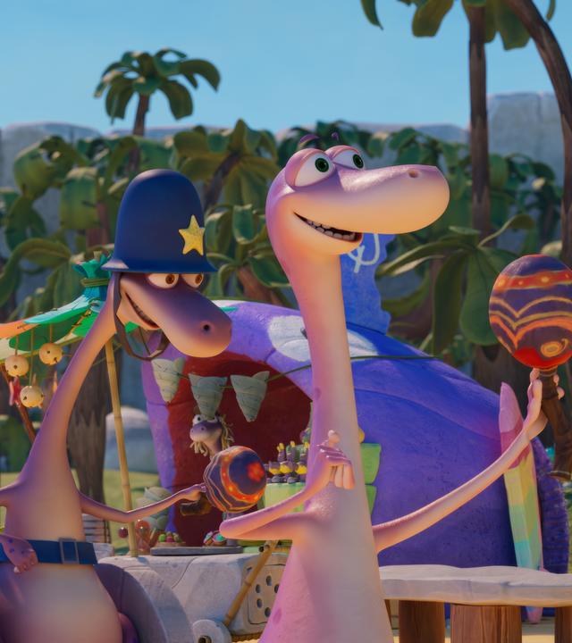 Der Dinosaurier Minus Drei hat eine farbenfrohe Rassel in der Hand. Hinter ihm rollt Wachtmeister Paleowski in seinem Rollstuhl. Er schwingt ebenfalls eine Rassel. Und noch weiter dahinter in der Reihe tanzen zwei mit Blumen geschmückte Dinosaurier. Sie scheinen eine Art Umzug zu machen und sind gutgelaunt.