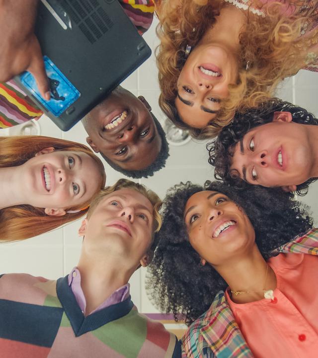 Blick von unten: Die Freunde haben sich Arm in Arm und Kopf an Kopf in einem Kreis aufgestellt und blicken nach unten Richtung Kamera: Mia, Jack, Simone, Benoit, Cece und Finn. Jack hält ein Laptop an den Oberkörper gepresst in den Armen. Alle tragen Freizeitkleidung und lächeln.