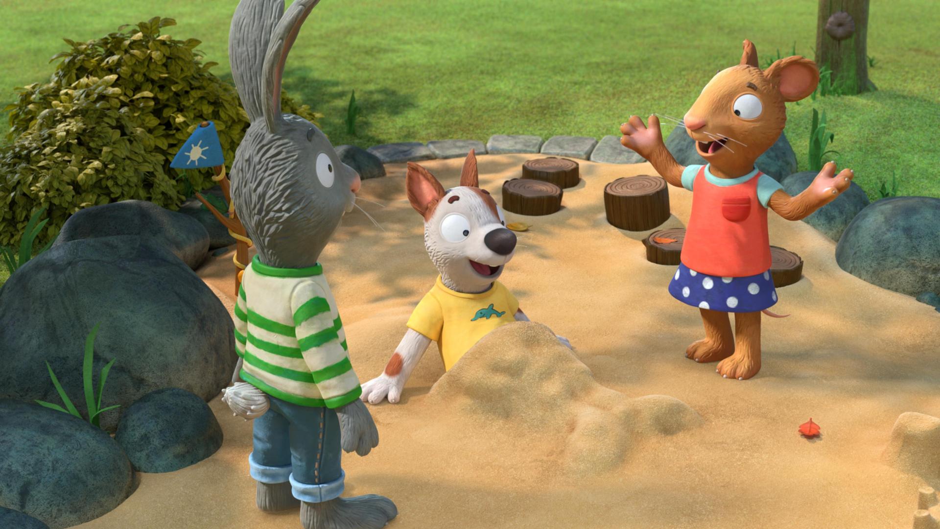 Auf dem Spielplatz im Sandkasten: Pip, das Kaninchen und Posy, die Maus freuen sich über den Anblick von ihrem Freund Zac. Der Hund Zac liegt im Sandkasten, hat sich halb aufgerichtet und blickt begeistert auf seine untere Hälfte, die komplett mit Sand bedeckt ist. Es hat die Form einer Meerjungfrauenflosse.