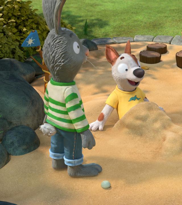 Auf dem Spielplatz im Sandkasten: Pip, das Kaninchen und Posy, die Maus freuen sich über den Anblick von ihrem Freund Zac. Der Hund Zac liegt im Sandkasten, hat sich halb aufgerichtet und blickt begeistert auf seine untere Hälfte, die komplett mit Sand bedeckt ist. Es hat die Form einer Meerjungfrauenflosse.