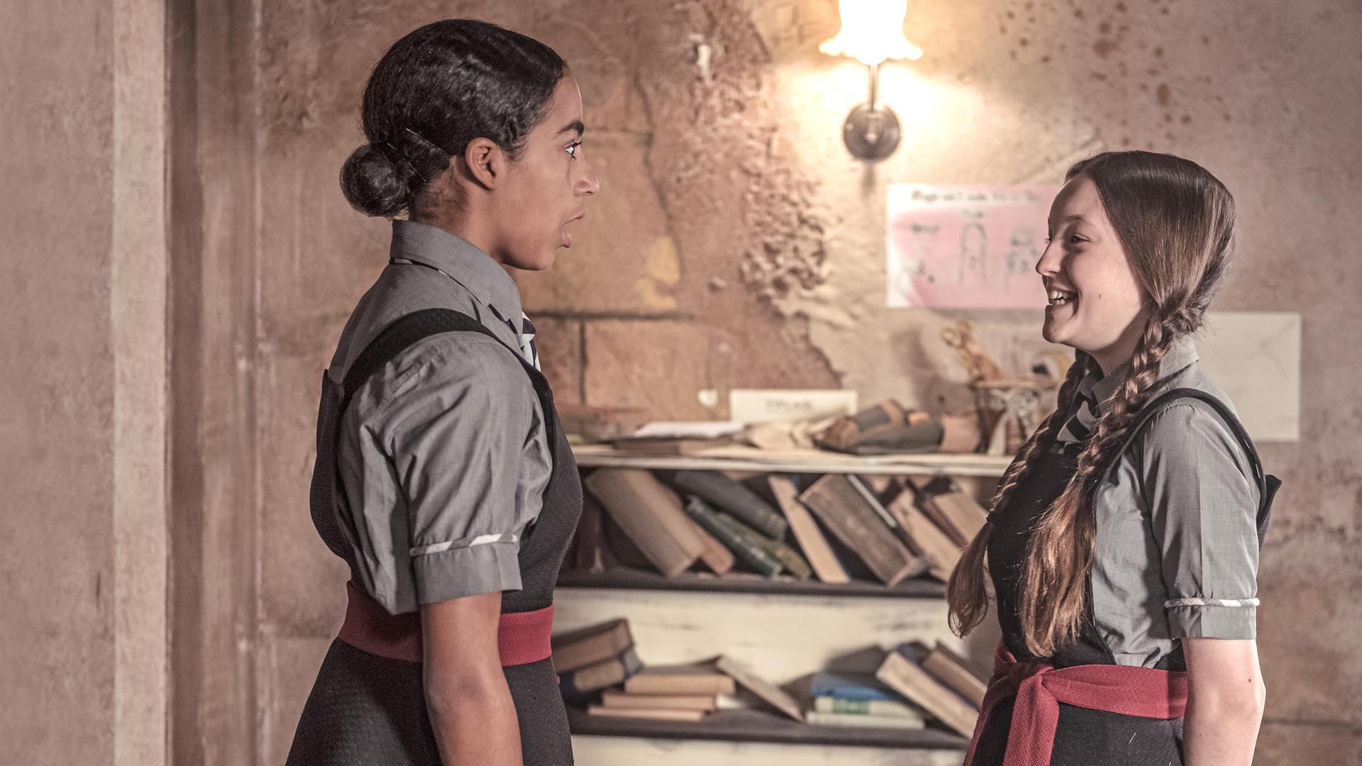 Mildred und Indigo stehen sich gegenüber und schauen sich an. Mildred lacht und Indigo schaut erstaunt.