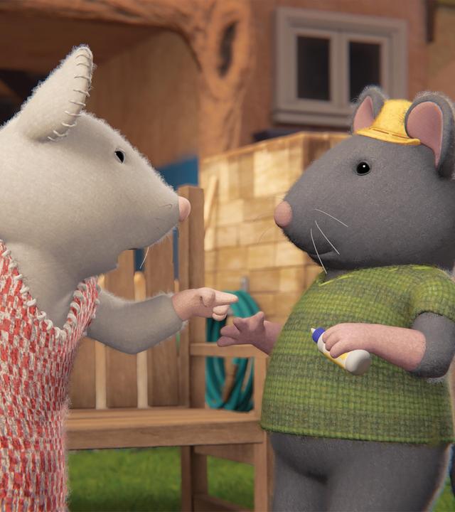 Julia steht vor Tonio, spricht zu ihm und deutet mit ihrem linken Zeigefinger auf ihn. Die beiden stehen im Garten vor einer Parkbank. Im Hintergrund ist die Fassade des Mäusehauses zu sehen.