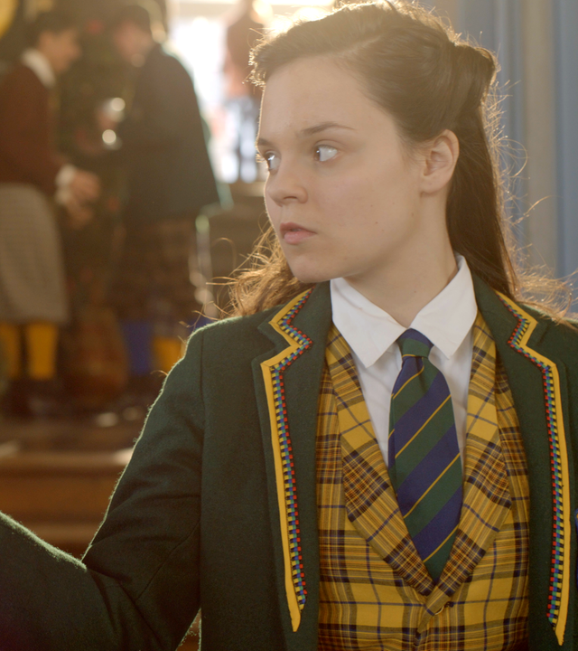 Theo wirkt angespann und hält einer Schülerin, die ihr den Rücken zukehrt, ein Amulett an den Rücken.