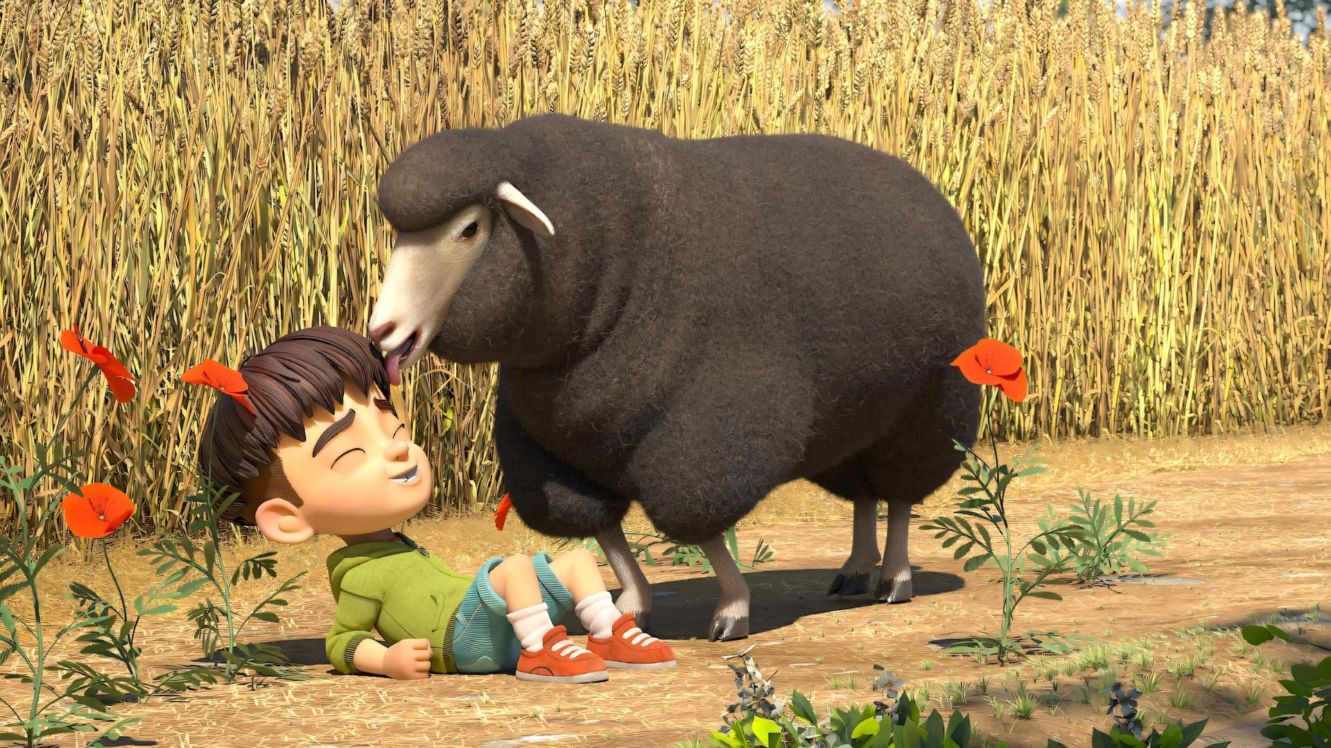 Ein großes, flauschiges und braunes Schaf leckt mit seiner Zunge über die Haare und das Gesicht von Kleiner Fuß. Dieser liegt lachend vor dem Schaf auf dem Feldboden vor einem reifen Kornfeld.