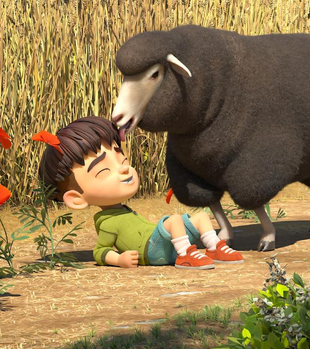 Ein großes, flauschiges und braunes Schaf leckt mit seiner Zunge über die Haare und das Gesicht von Kleiner Fuß. Dieser liegt lachend vor dem Schaf auf dem Feldboden vor einem reifen Kornfeld.