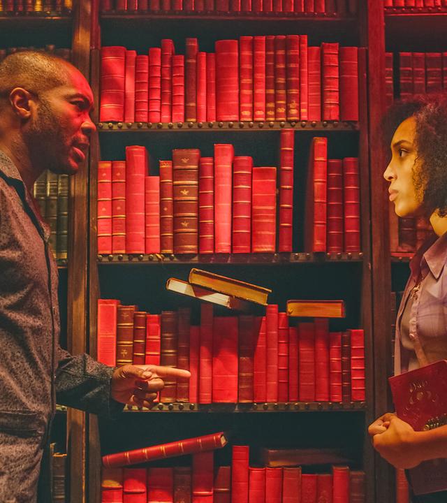 In einer dunklen, nur spärlich beleuchteten Bibliothek vor Regalen voller Bücher steht Cece ihrem Vater David Parker gegenüber. Die Stimmung wirkt angespannt. David zeigt mit dem Finger auf Cece und spricht mit ihr. Cece blickt ihn ernst an und hat dabei nervös ihre Hände ineinander verschränkt. Sie hält ein Buch eng an ihren Oberkörper gedrückt.