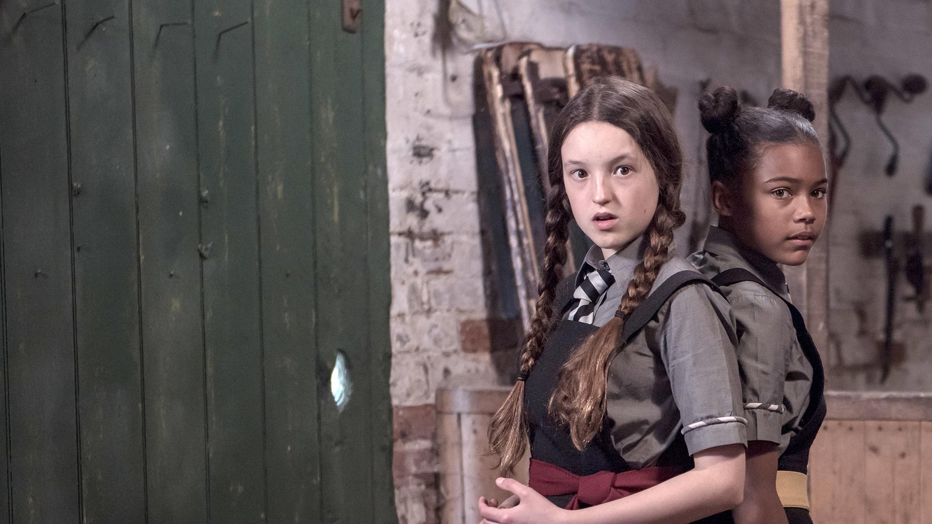 Mildred und Edith stehen Rücken an Rücken in einer Besenkammer. Sie schauen bestürzt auf etwas.