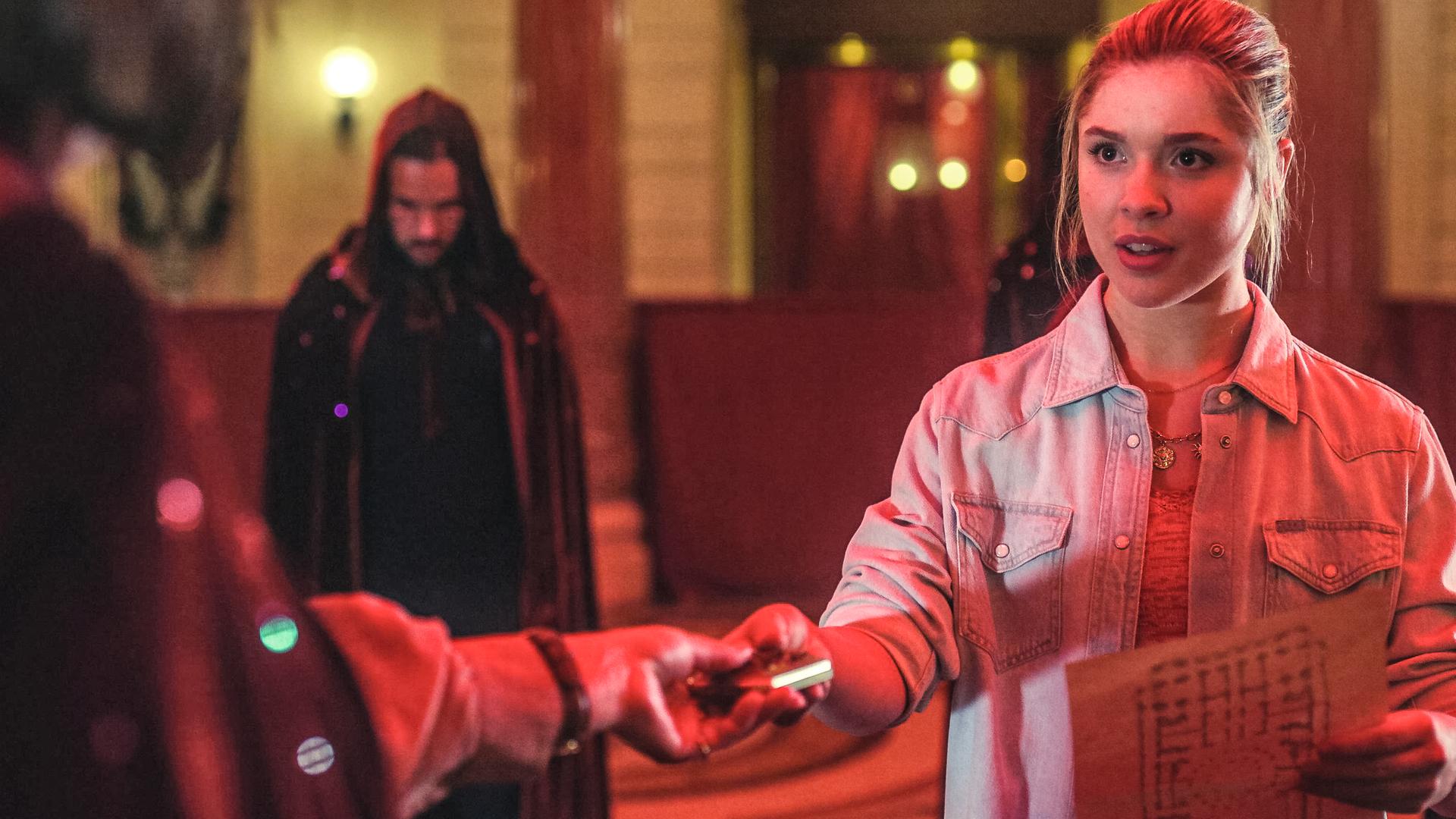 Jade hält einen Lageplan in ihrer linken Hand. Sie reicht ihre Mutter Zoe, die im Bild nur im Anschnitt und von hinten zu sehen ist, einen Gegenstand. Im Hintergrund stehen Männer des Mystik-Zirkels. Sie tragen Umhänge mit Kapuzen, die sie über den Kopf gezogen haben.