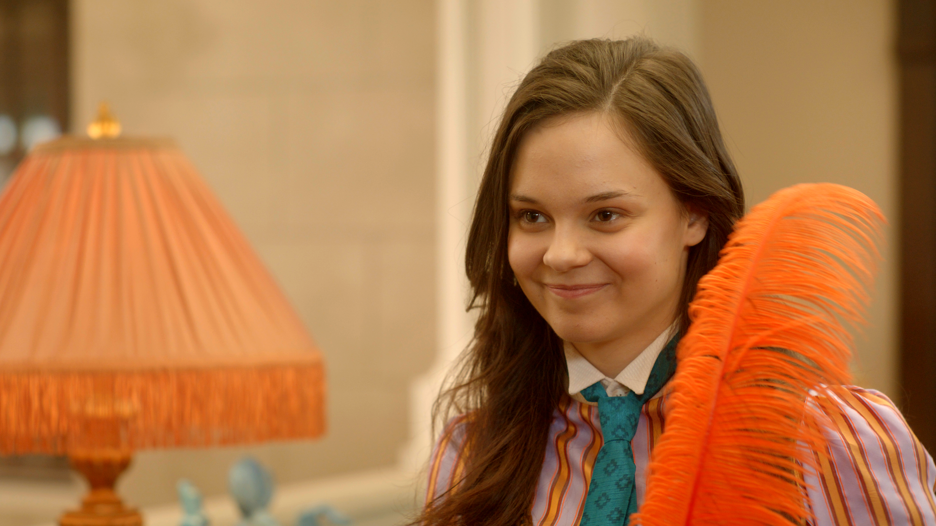 Theo hält eine orange eingefärbte Straußenfeder in ihren Händen. Sie lächelt glücklich. Neben ihr auf einer Kommode steht eine Leuchte mit einem ähnlich orangefarbenen Lampenschirm.