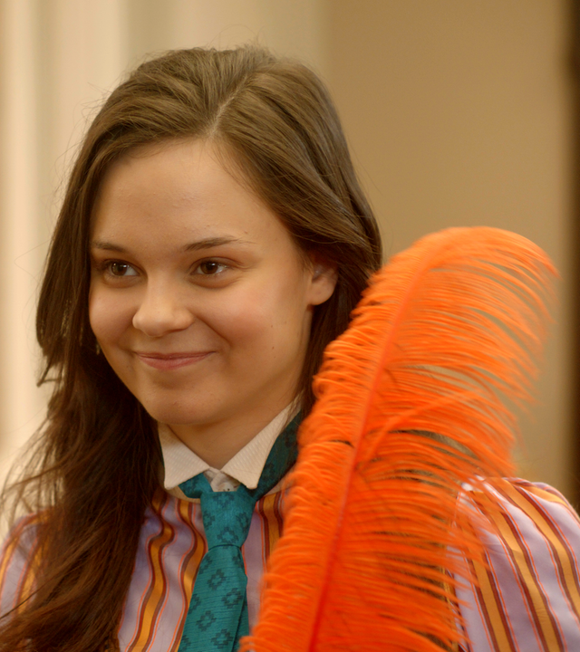 Theo hält eine orange eingefärbte Straußenfeder in ihren Händen. Sie lächelt glücklich. Neben ihr auf einer Kommode steht eine Leuchte mit einem ähnlich orangefarbenen Lampenschirm.