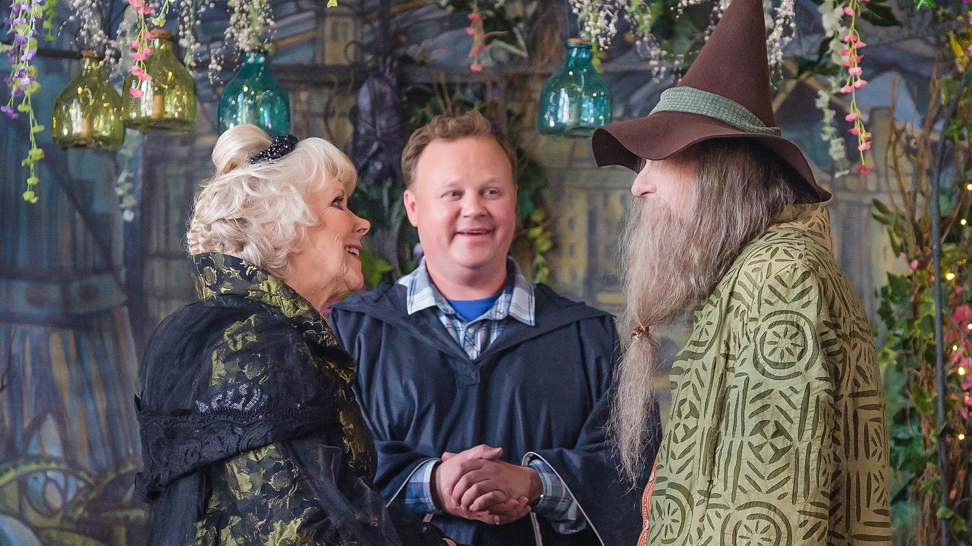 Frau Maus und Herr Braunweber stehen vor einem Mann am Altar. Um sie herum sind überall Blumengirlanden.