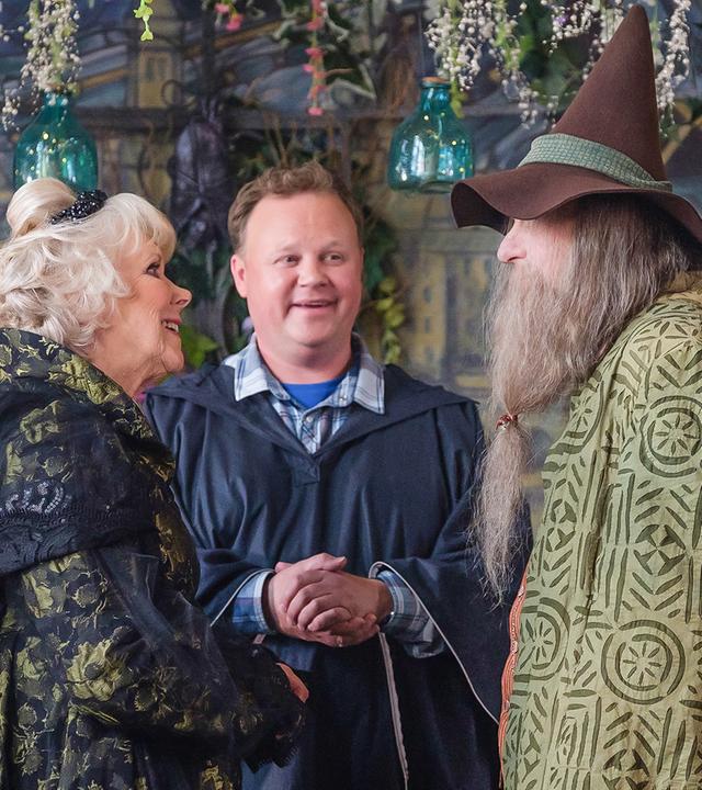 Frau Maus und Herr Braunweber stehen vor einem Mann am Altar. Um sie herum sind überall Blumengirlanden.