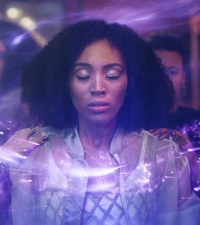 Cece steht mit geschlossenen Augen in einem Raum voller älterer Menschen. Es sind die Wizen-Ältesten, die sie erwartungsvoll ansehen. Manche haben ihre Hände auf Ceces Schultern abgelegt. Um Cece herum schweben blau-violett schimmernde Lichter. Cece sieht angestrengt aus.