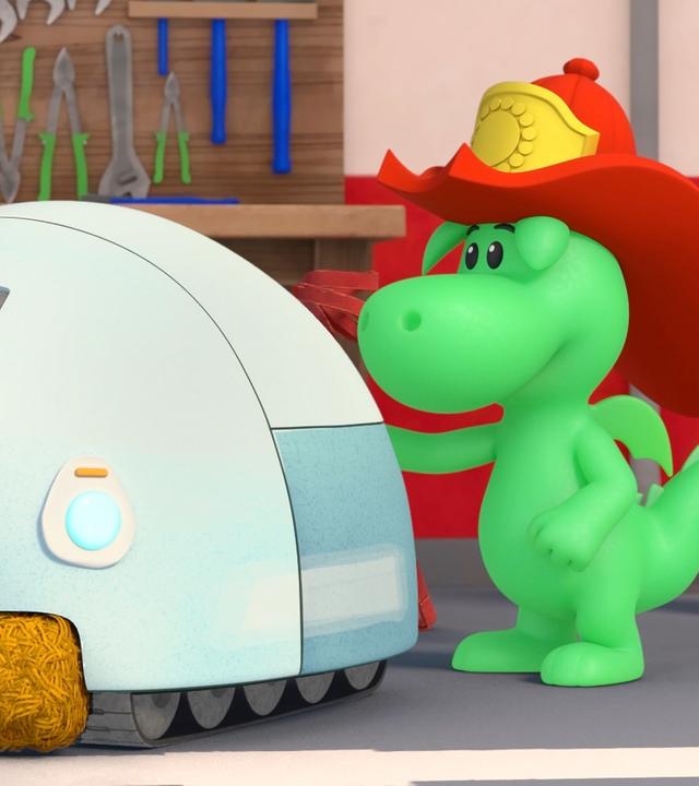 Grisu, der kleine, grüne Drache mit den roten Feuerwehrhelm, seine Freundin Stella und Hund Dinki stehen um einen Roboter herum, der aussieht wie ein runder, großer Mähroboter.