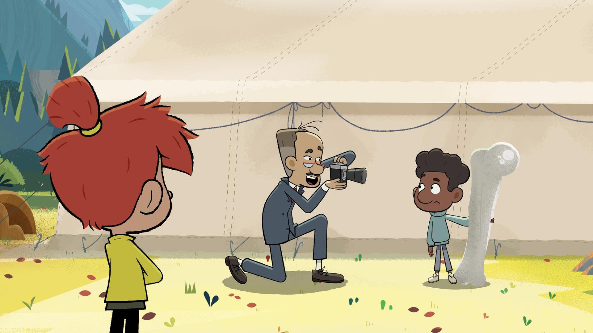 Tommy steht mit dem großen Knochen in der Hand neben einem großen Zelt. Vor ihm kniet ein Mann, der ihn fotografiert. Taylor steht im Vordergrund und schaut dabei zu.