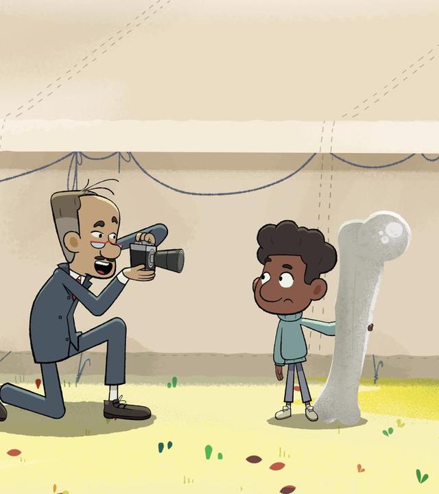 Tommy steht mit dem großen Knochen in der Hand neben einem großen Zelt. Vor ihm kniet ein Mann, der ihn fotografiert. Taylor steht im Vordergrund und schaut dabei zu.