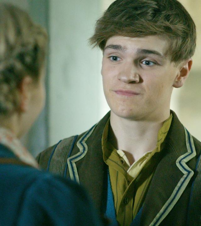 Henry schaut etwas verzweifelt auf Maya, von der wir nur den Rücken und ihre geflochtenen Haaren sehen.