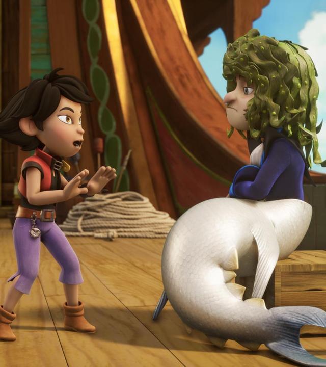 Käpt'n Flint sitzt als Meerjungfrau verkleidet auf einer Holzkiste. Er hat eine Haiflosse an und Meeralgen als Perücke auf seinem Kopf. Vor ihm steht Zoe und hält in einer Abwehrhaltung mit ihrem Händen nach vorne.