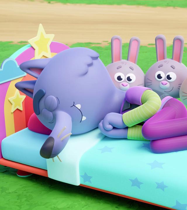 In einem Bettchen liegt Wilf, der lilafarbene Wolf mit Hose und Badelatschen. Er schläft tief und fest. Hinter dem Bett stehen sechs kleine Häschen.