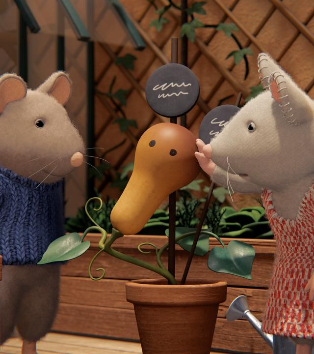 Sam und Julia stehen auf der Terrasse zwischen Hochbeeten und betrachten von beiden Seiten einen Kürbis, der in einem Topf gewachsen ist. Er hat zwei runde Flecken, die wie Augen aussehen, und Julia tätschelt liebevoll den Kürbis-Kopf.