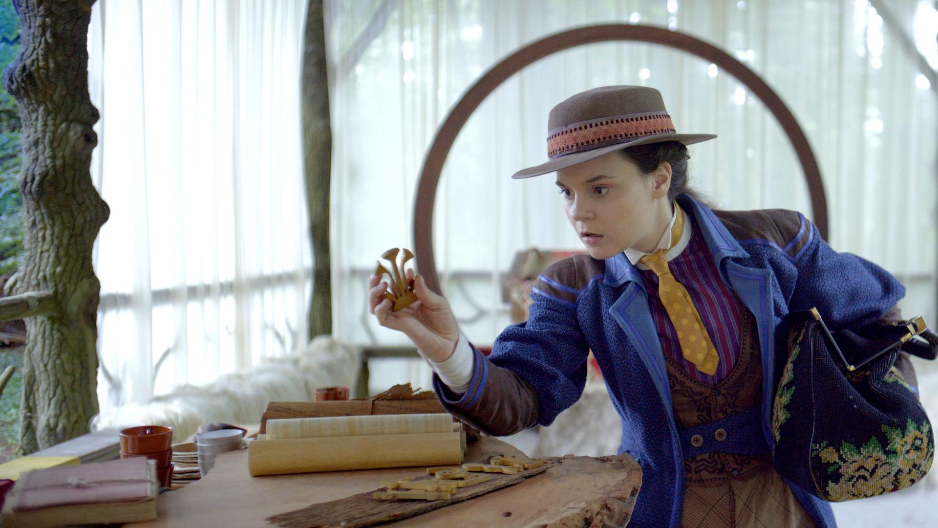 Auf der Terrasse vom Refugium der Schwesternschaft steht Theodosia, die sich über einen Tisch beugt, der aus einer Baumstammplatte besteht. Auf diesem Tisch liegen mehrere Gegenstände sowie Pergamentrollen. In ihrer rechten Hand hält sie nachdenklich einen handtellergroßen Gegenstand. Unter ihrem linken Arm trägt sie eine große, weit geöffnete Tasche. Verschwommen im Hintergrund der Terrasse, die teils mit weißen Vorhängen zugehängt ist, ist ein Waldstück zu erkennen.