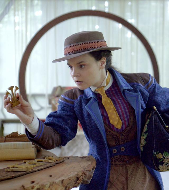 Auf der Terrasse vom Refugium der Schwesternschaft steht Theodosia, die sich über einen Tisch beugt, der aus einer Baumstammplatte besteht. Auf diesem Tisch liegen mehrere Gegenstände sowie Pergamentrollen. In ihrer rechten Hand hält sie nachdenklich einen handtellergroßen Gegenstand. Unter ihrem linken Arm trägt sie eine große, weit geöffnete Tasche. Verschwommen im Hintergrund der Terrasse, die teils mit weißen Vorhängen zugehängt ist, ist ein Waldstück zu erkennen.